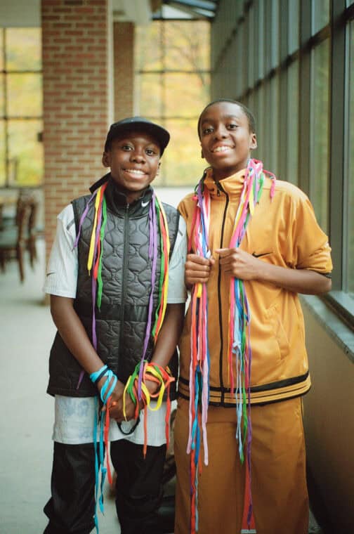 Two young people smiling at the camera with many shoelaces in different colours hanging around their necks.