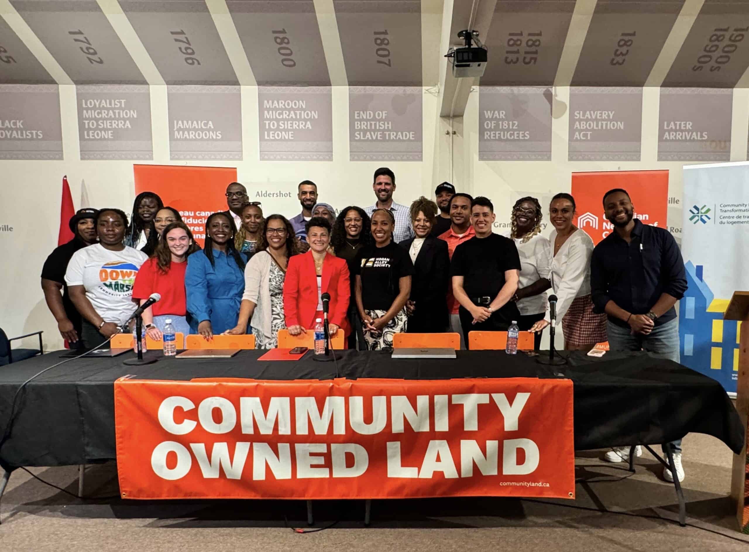 A group of 22 people stand behind a table that says "Community Owned Land"
