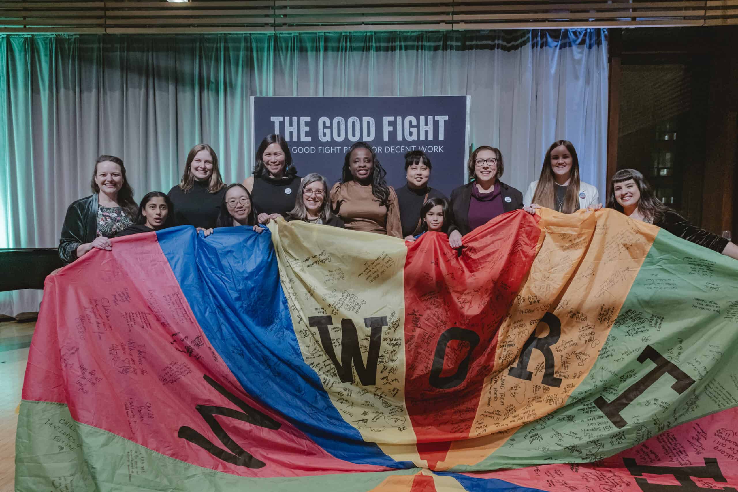 12 people smiling and posing in front of a sign that reads "The Good Fight Prize for Decent Work" and hold a colourful parachute that has letters and signatures or small writing on it.