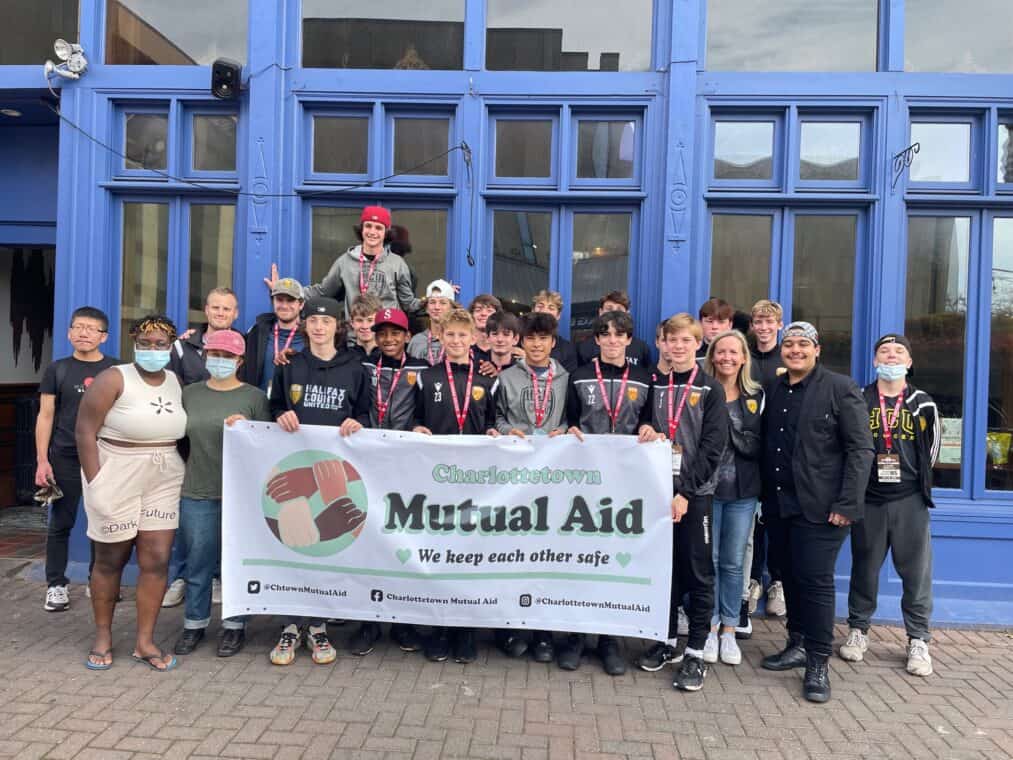 Large group of people posing in front of a building with a sign that reads, "Charlottetown Mutual Aid: We keep each other safe"