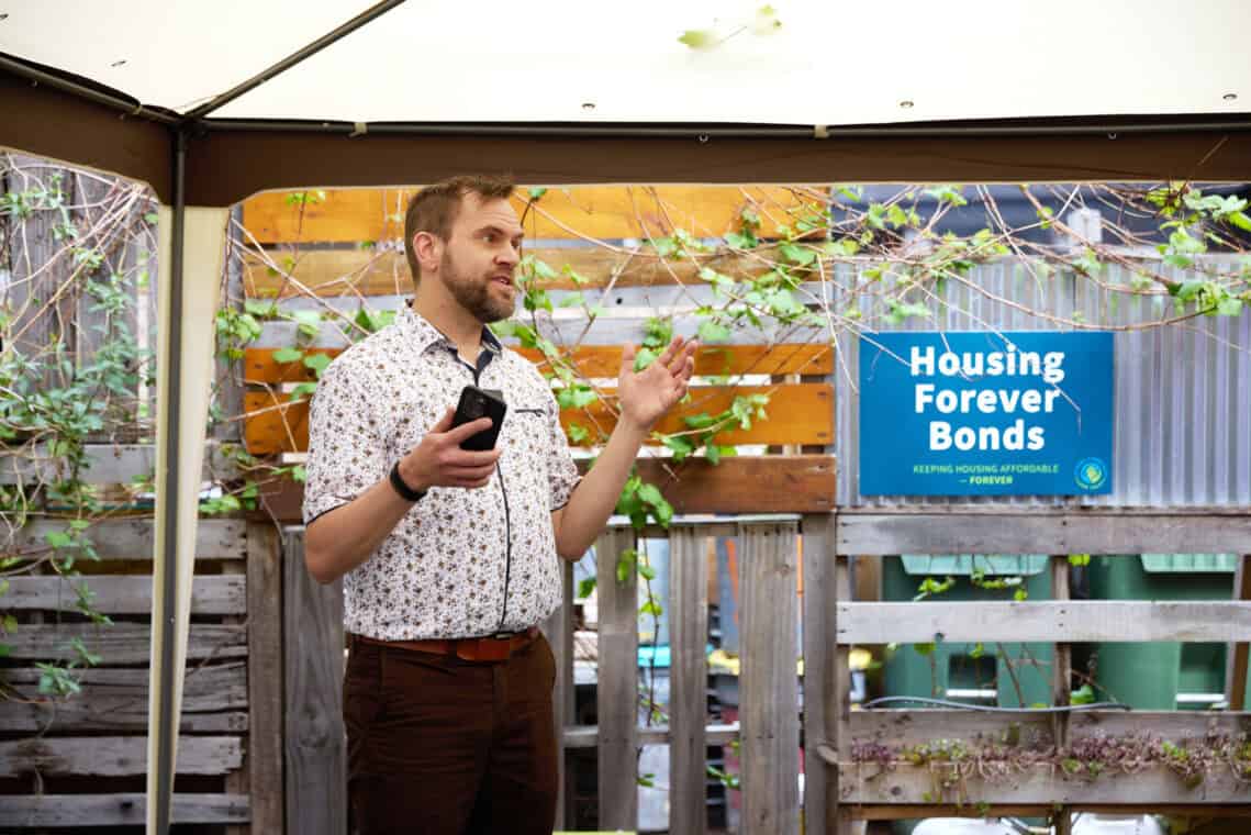 Person speaking under a garden tent. There is a sign in the background that says "Housing Forever Bonds"