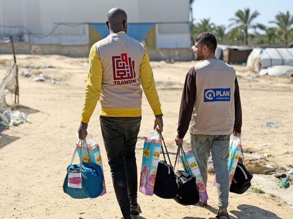 Two people in humanitarian vests are carrying bags of supplies.