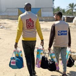 Two people in humanitarian vests are carrying bags of supplies.