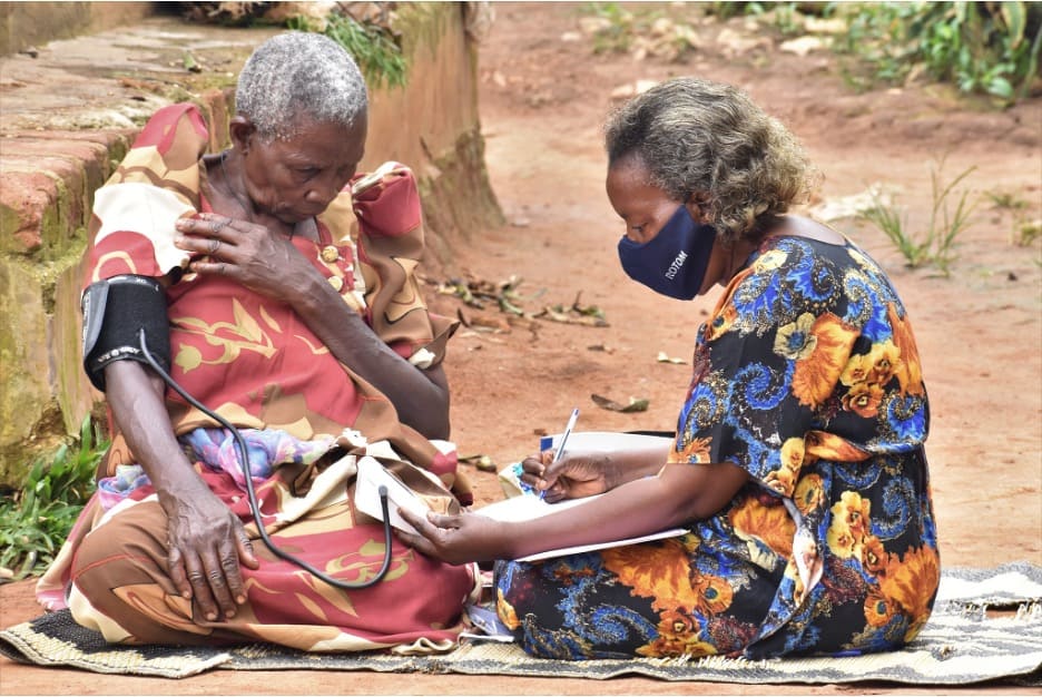 Two people sitting on a mat outdoors while one person checks the blood pressure of another and records something in a notebook.