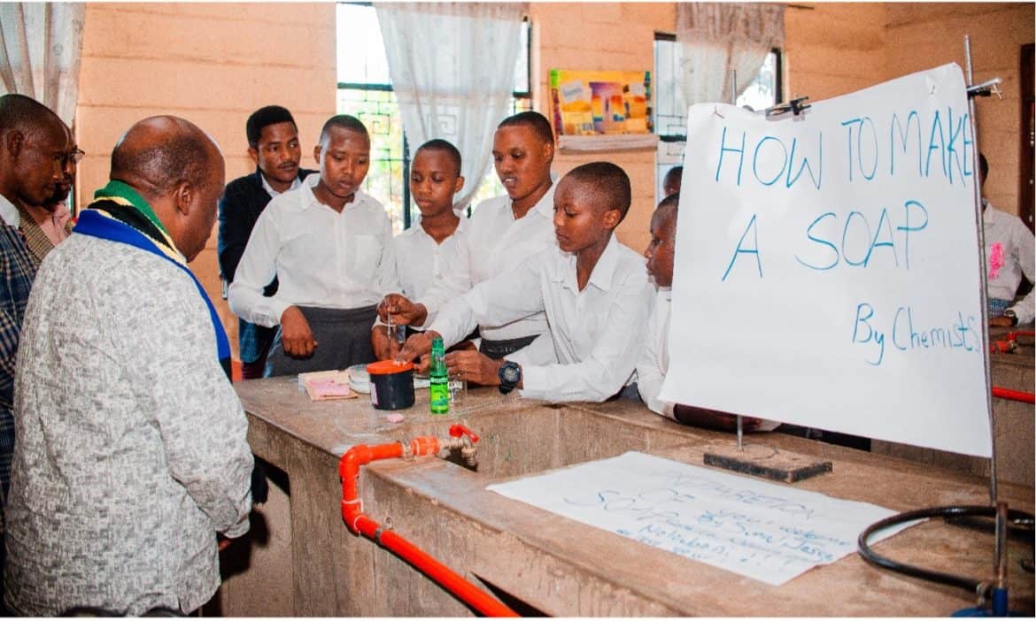 Several students in uniforms surround science materials. There is a handwritten sign that says "how to make soap."