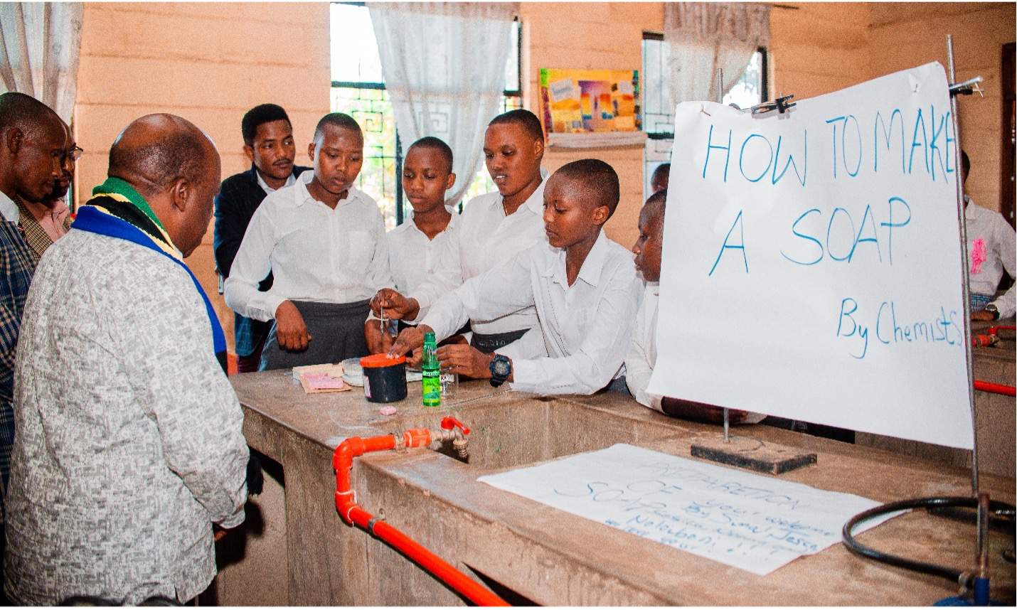 Several students in uniforms surround science materials. There is a handwritten sign that says "how to make soap."