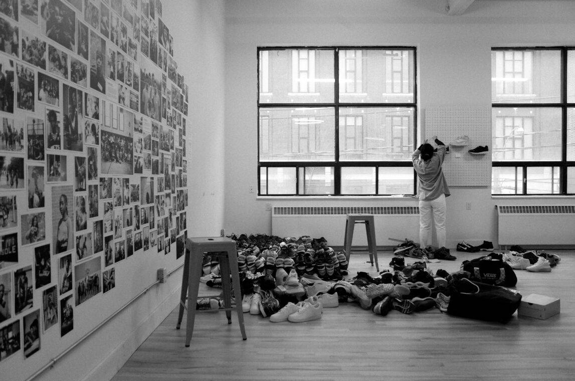 A big room with pictures covering the wall to the left. Piles of shoes on the floor. Someone is placing a few of the shoes on a display wall.