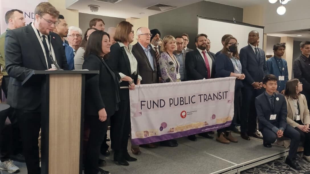 Large group of people in suits surround a banner that reads "fund public transit"
