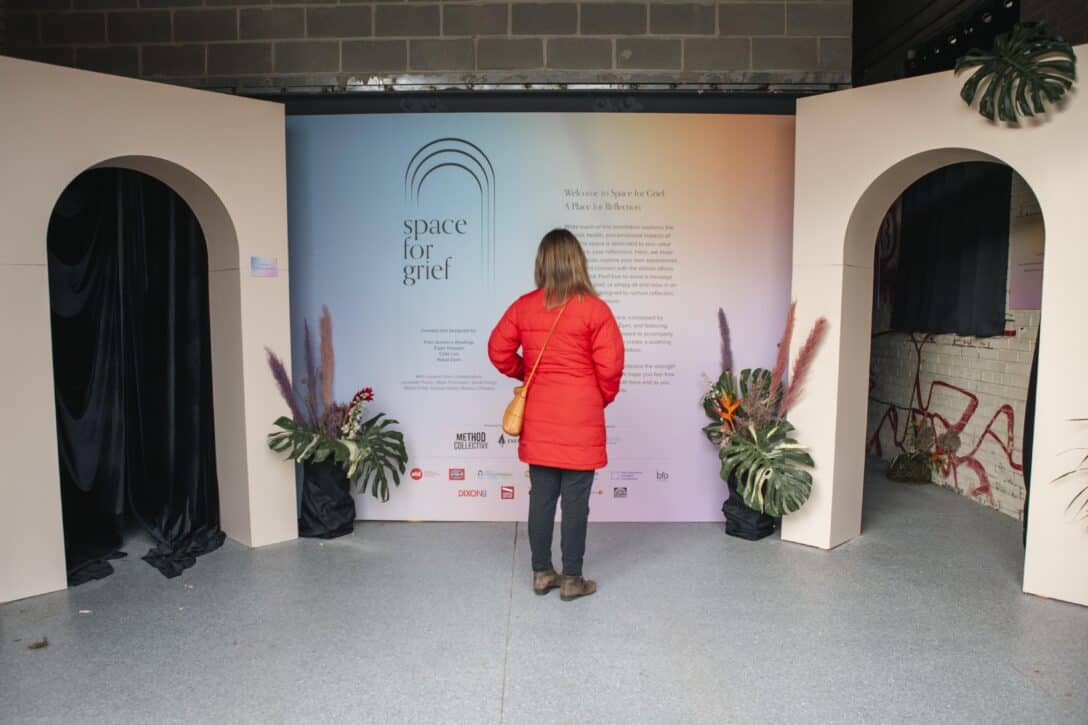 A woman in a red jacket stands alone reading about the Space for Grief project, surrounded by plants between the arched entry and exits of the installation.