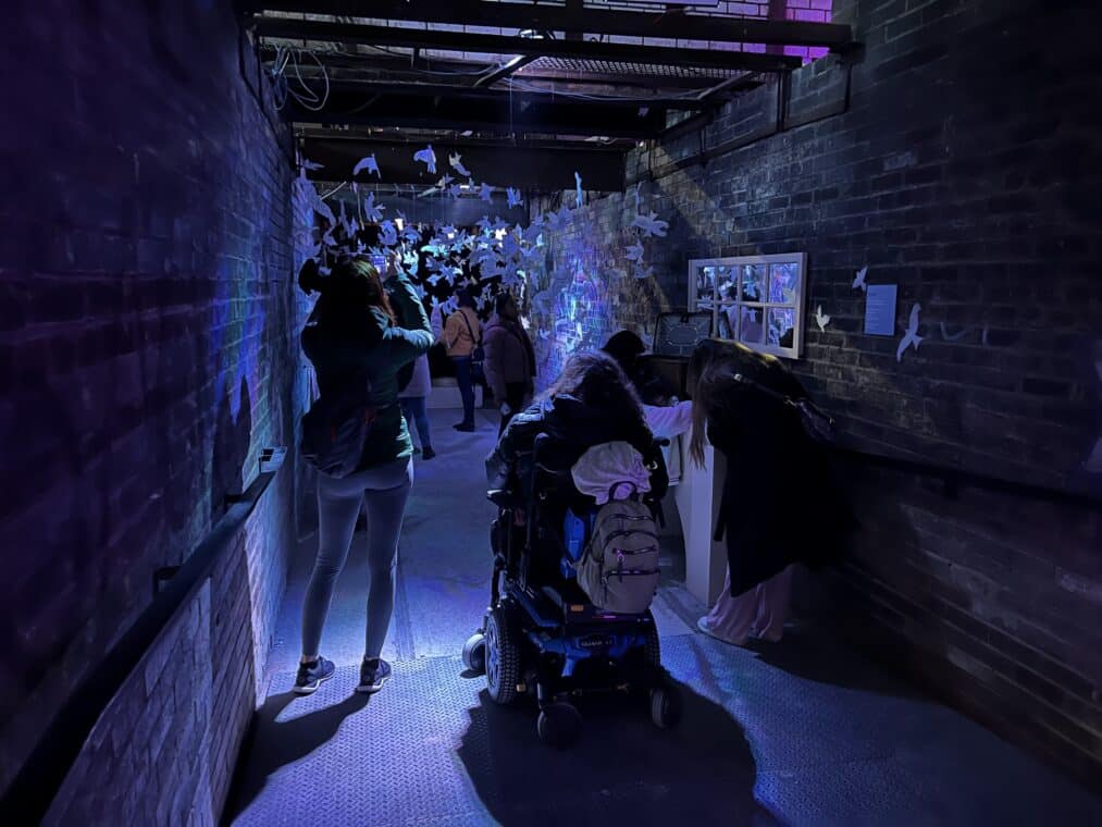A group of people gathered in a hallway lightly lit with streams of blue and purple. Paper birds hang from the ceiling carrying people’s messages to their grief. Some people are standing taking photos, another in a wheelchair, and a few at a podium on the side writing their messages to be hung.