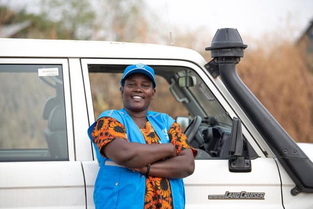 A person leaning against a car with their arms folded wearing a blue vest and hat with the UNHCR symbol.