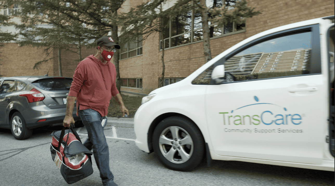 Person carrying an insulated bag and walking to a car that says "TransCare Community Support Services" on the door.